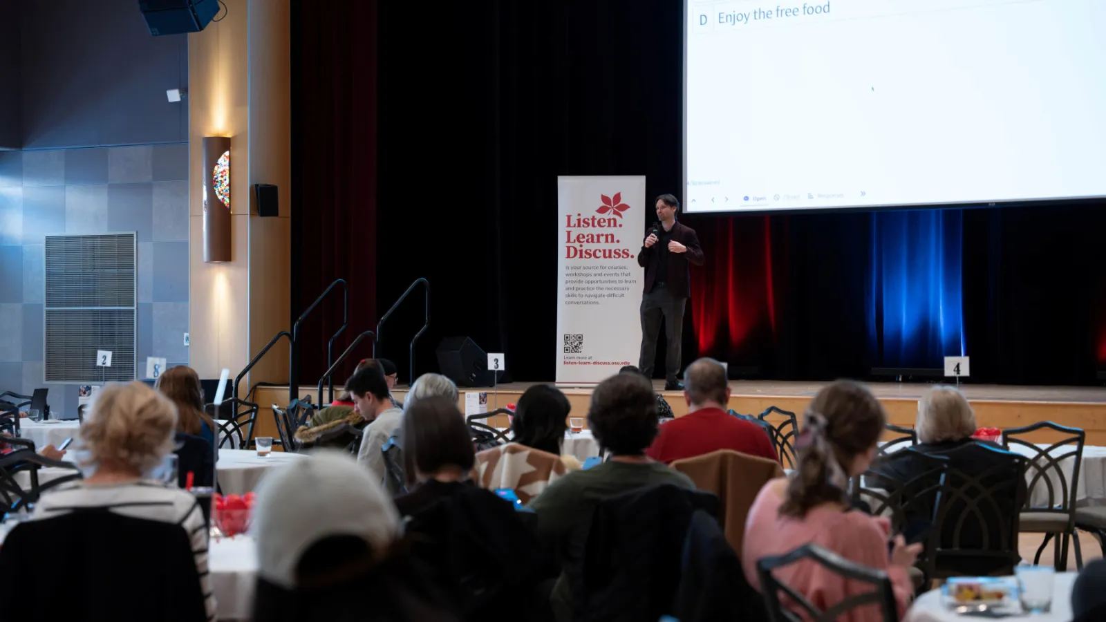 Aaron Yarmel stands on a stage talking to participants at round tables